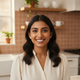 Portrait of a smiling South Asian woman looking at the viewer, set against a softly blurred kitchen background in warm tones.